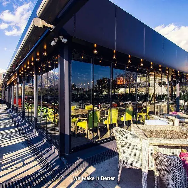 کافه کارزین با فضای مدرن شیشه ای، سقف متحرک، و تراس دنج دارای صندلی های حصیری و منظره کوهستانی در روز آفتابی.