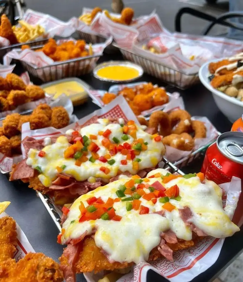 میز پر از غذاهای فست فود قلب طلایی نیاوران شامل ساندویچ‌های لذیذ با پنیر ذوب شده و فلفل، ناگت مرغ، سیب‌زمینی و پیاز سوخاری.