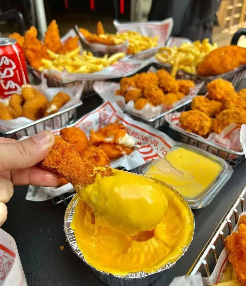 یک دست در حال فرو بردن چیکن استریپس طلایی در سس پنیر لذیذ در فست فود قلب طلایی نیاوران، همراه با سیب زمینی و ناگت.