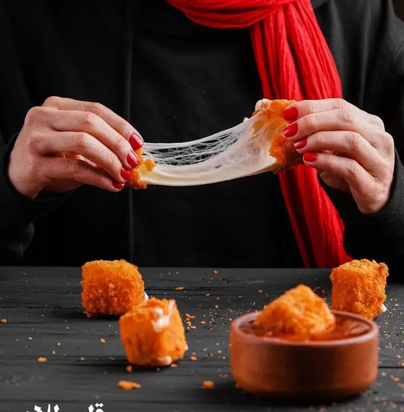 تصویری جذاب از کش آمدن پنیر داغ از لقمه سوخاری فست فود قلب طلایی نیاوران، با تکه‌های خوشمزه دیگر روی میز.