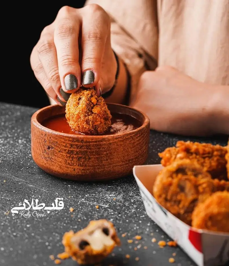 دستی در حال فرو بردن لقمه سوخاری پنیردار ترد در سس قرمز. بهترین و کریسپی‌ترین غذاها را از فست فود قلب طلایی نیاوران بخواهید.