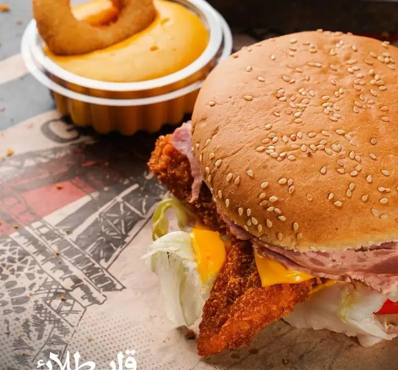 عکس برگر سوخاری تازه و خوشمزه از فست فود قلب طلایی نیاوران، با نان کنجدی، کاهو، پنیر و سس دلپذیر، در کنار پیاز حلقه ای.