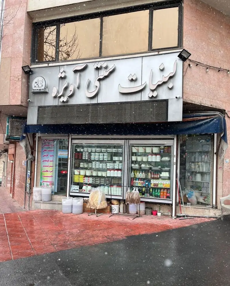 نمای بیرونی فروشگاه لبنیات سنتی آویژه (یزدی) در برف ملایم، با ویترین پر از ماست، شیر، پنیر و سایر محصولات لبنی تازه و باکیفیت.