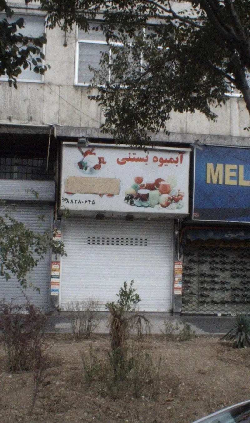 عکس یک مغازه آبمیوه بستنی با تابلوی جذاب شامل میوه و نوشیدنی‌ها، کرکره بسته فلزی و درختان اطراف آن.