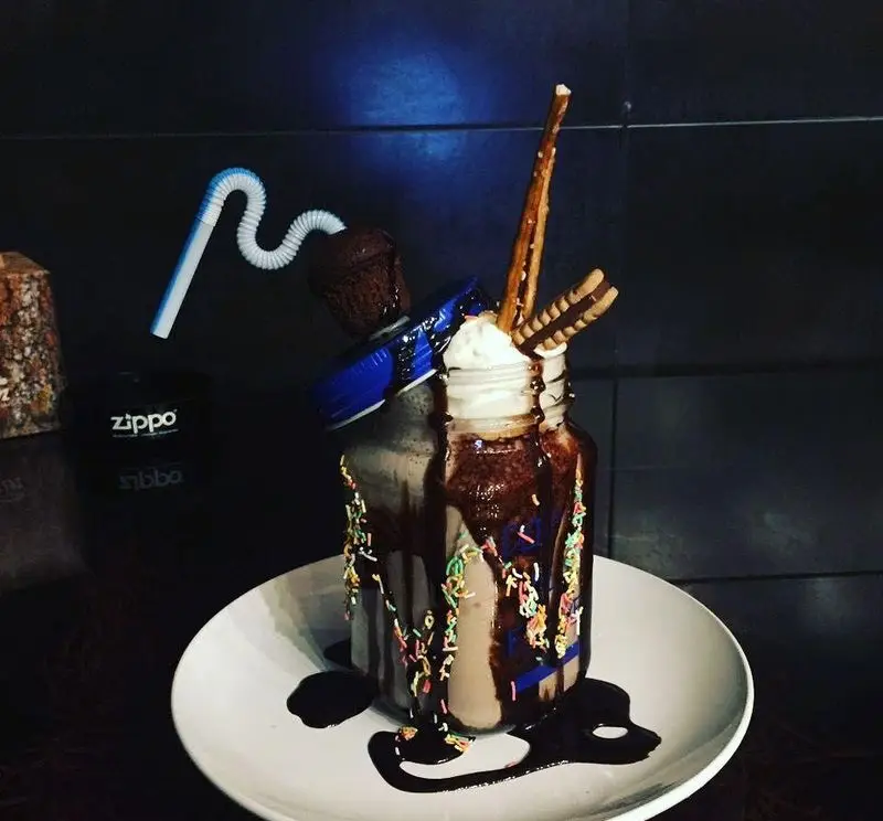 میلکشیک دبل شکلاتی ویژه کافه رورا با تزیینات جذاب پرینکل، سس شکلات، خامه، کاپ کیک و چوب شور در لیوان شیشه‌ای.