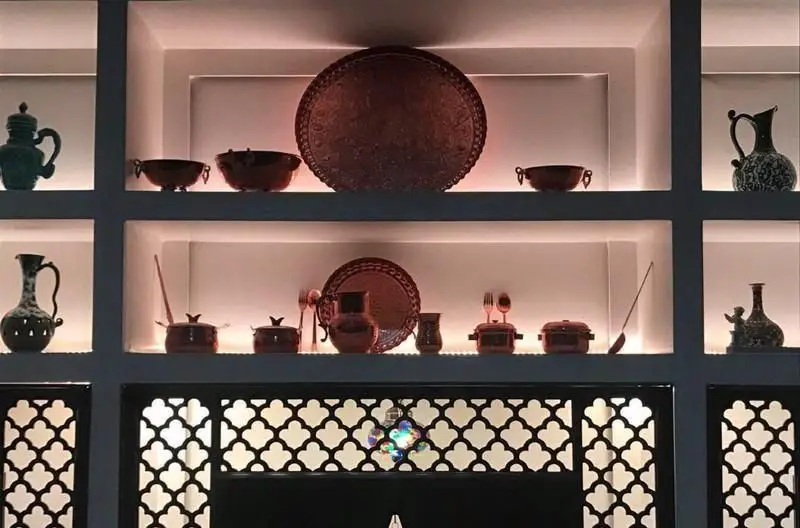 نمایی از دکوراسیون داخلی دلنشین رستوران رستاک، با قفسه‌های سنتی نورانی، ظروف مسی و طرح‌های هنری ایرانی در محیطی آرامش‌بخش.