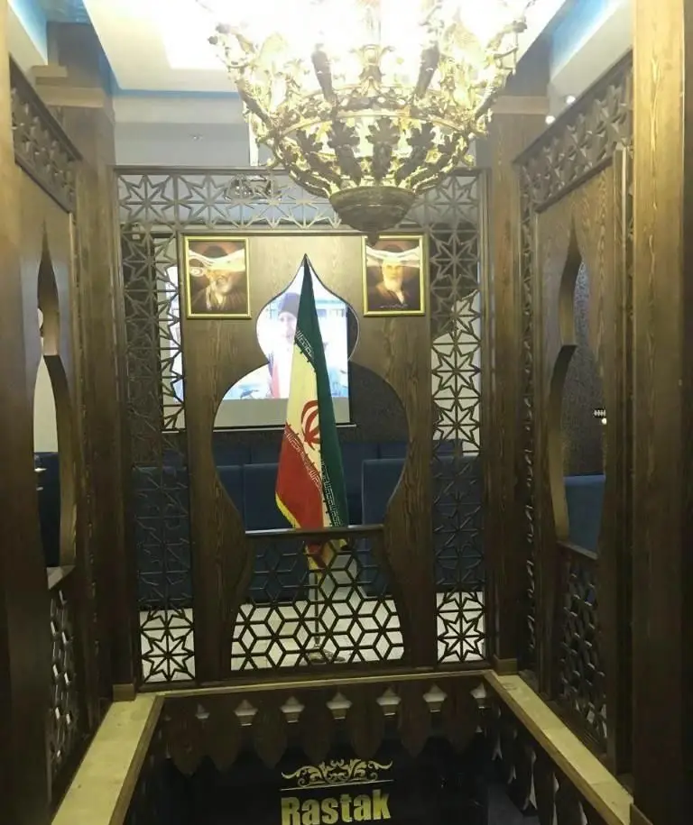 فضای داخلی رستوران رستاک با لوستر باشکوه، طرح‌های چوبی اسلامی و پرچم ایران، نمایانگر فضایی سنتی و دلنشین ایرانی.