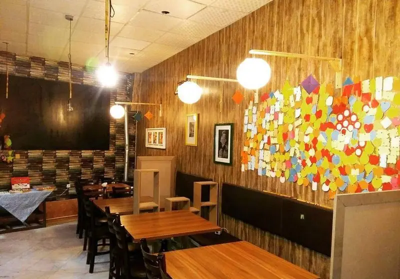 فضای داخلی کافه رستوران دنج با دکوراسیون گرم، میز و صندلی چوبی، دیوارپوش طرح چوب و کلاژ رنگارنگ جذاب.