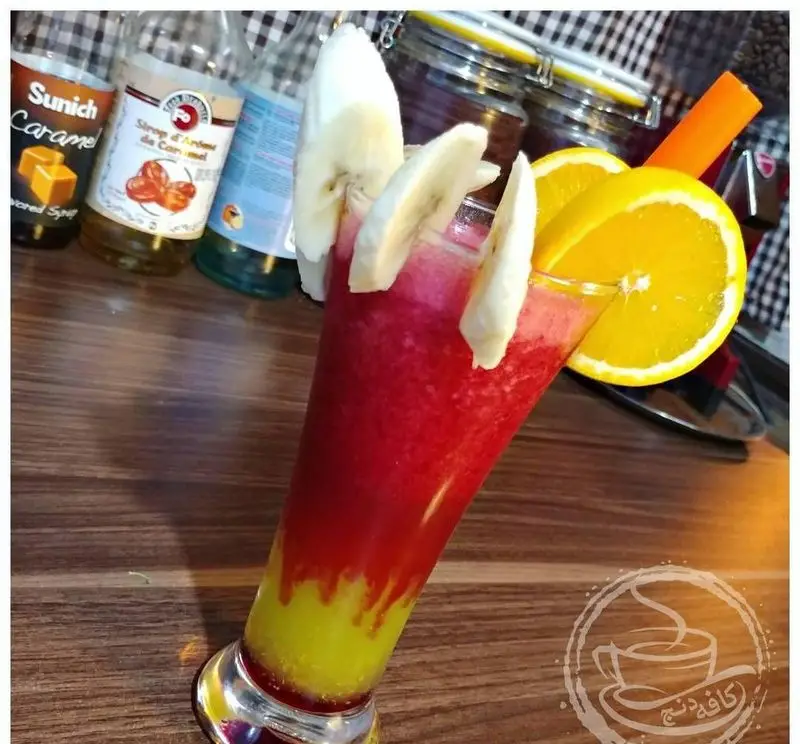 اسموتی میوه ای و طبیعی کافه رستوران دنج با لایه های رنگی جذاب و تزئین موز، پرتقال و هویج، نوشیدنی خنک و سالم.