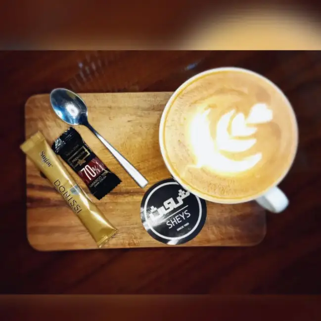 تصویری از یک فنجان قهوه لاته با طرح لاته آرت زیبا، همراه با شکر دونیسی و شکلات تلخ در کافی شاپ شیث (Sheys Coffee Shop). سرو نوشیدنی گرم.