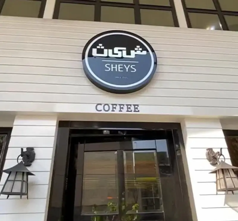 نمای بیرونی کافی شاپ شیث با تابلوی دایره‌ای شکل Sheys و کلمه Coffee بالای ورودی. معماری مدرن با سایدینگ سفید.