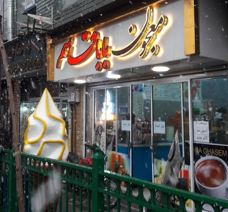 نمای خارجی مغازه معجون باباقاسم در روز برفی با تابلوی روشن، بستنی قیفی تزئینی بزرگ و ویترین شلوغ فروشگاه دسر و نوشیدنی.