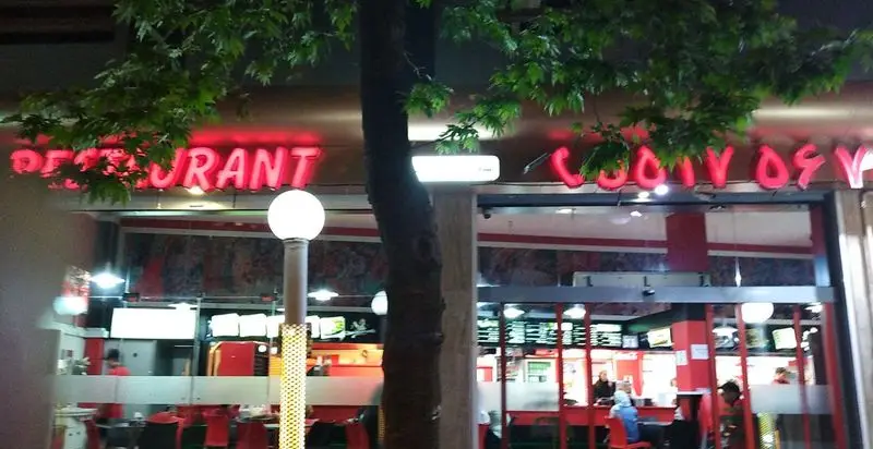 نمای شبانه رستوران جوجه بریانک با تابلوی نئون قرمز رنگ شامل عبارت 'RESTAURANT' و شماره تماس و نمای داخلی با مشتریان.