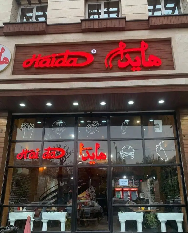 فست فود هایدا ده حقی، نمای بیرونی مدرن و جذاب با تابلوی درخشان فارسی و انگلیسی، همراه با نقاشی‌های فست فود روی شیشه‌ها.
