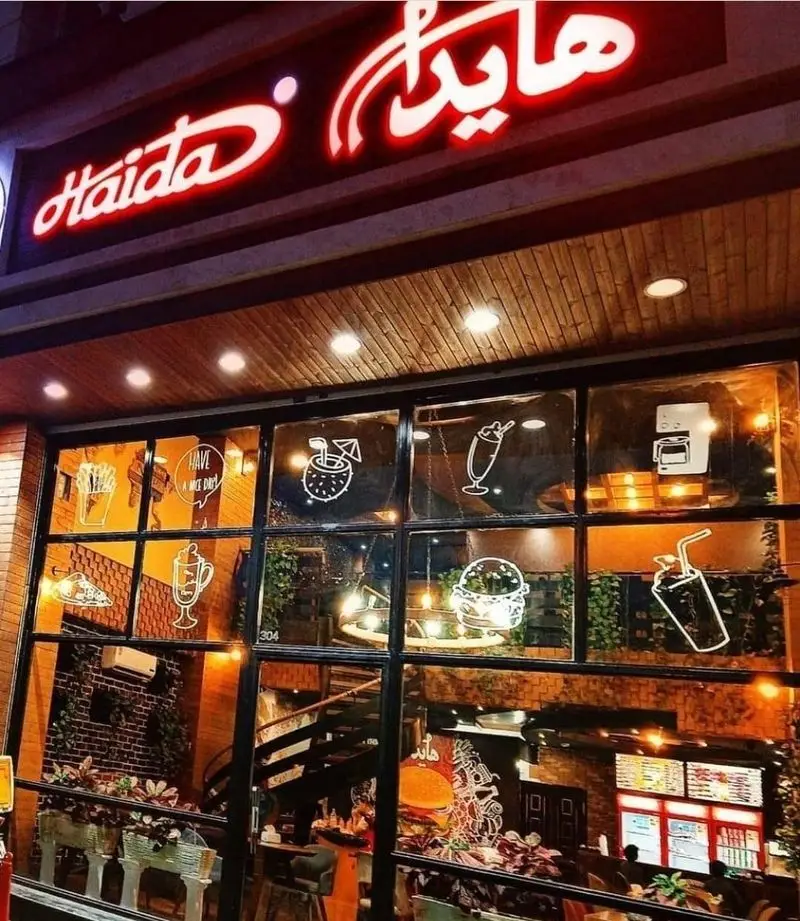 ورودی رستوران فست فود هایدا (Haida) شعبه ده حقی در شب، با تابلوی نئون قرمز و ویترین شیشه‌ای شامل طرح سیب‌زمینی، برگر، پیتزا و نوشیدنی‌های متنوع.