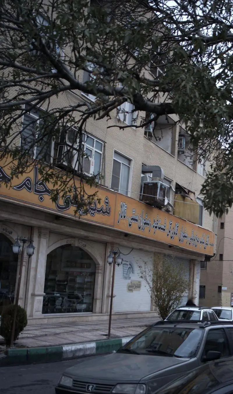 نمای بیرونی کسب و کار شیرینی جام جم، قنادی و رستوران سنتی ایرانی، با تابلوی زرد رنگ. خودروها در خیابان جلوی مغازه.