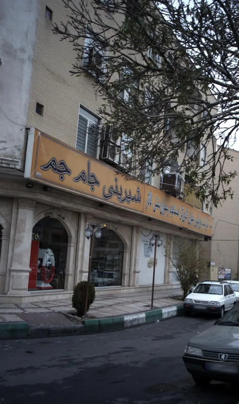 تصویری از نمای بیرونی شیرینی جام جم، قنادی و فروشگاه معروف شیرینی‌جات در خیابانی شلوغ در ایران، با ویترین‌های بزرگ، معماری کلاسیک و درختان.