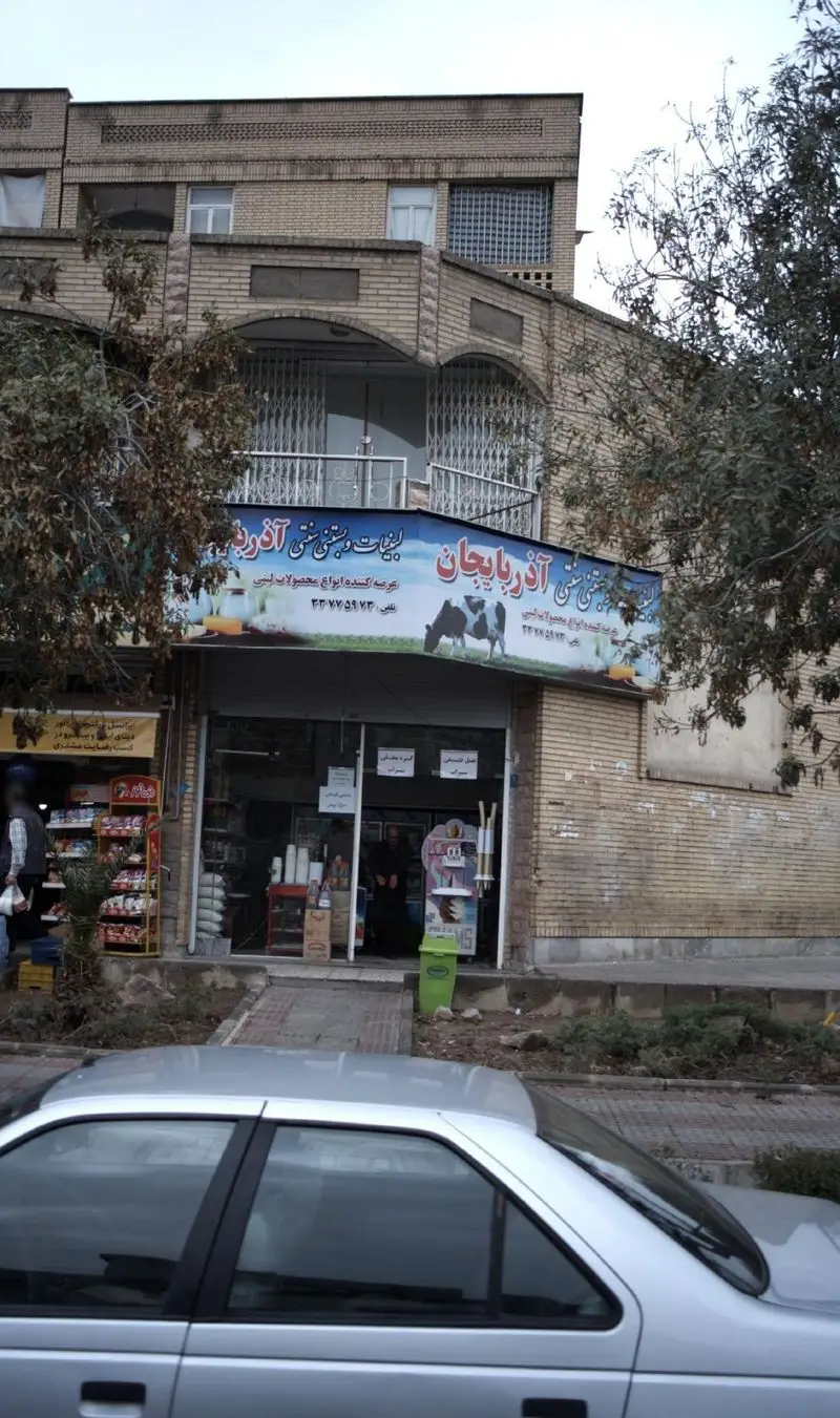 نمای خارجی مغازه لبنیات و بستنی سنتی آذربایجان با عرضه محصولات لبنی تازه، یک خودرو نقره‌ای و ساختمان آجری در پس‌زمینه
