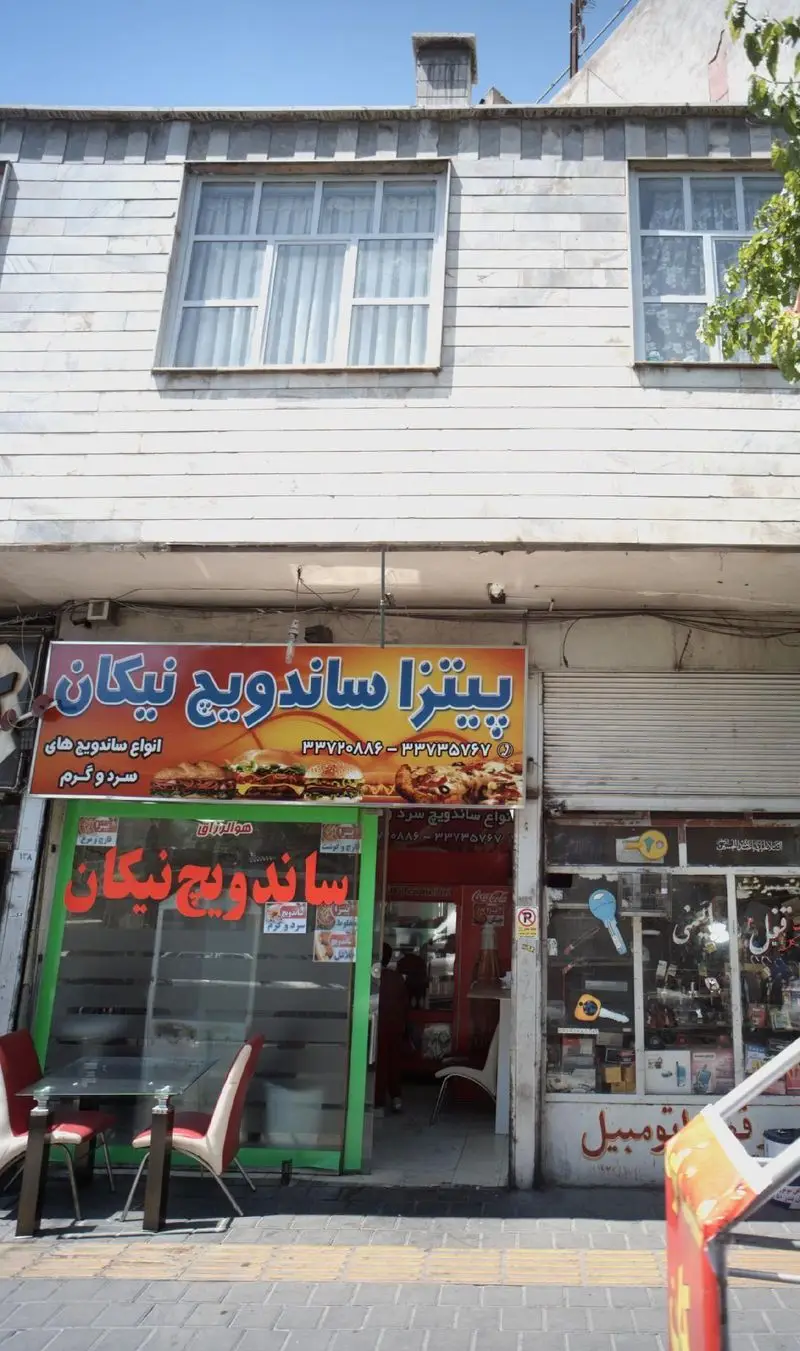 تصویر نمای جلوی مغازه پیتزا و ساندویچ نیکان. تابلوی بزرگ با تبلیغ انواع پیتزا، همبرگر و ساندویچ‌های سرد و گرم.