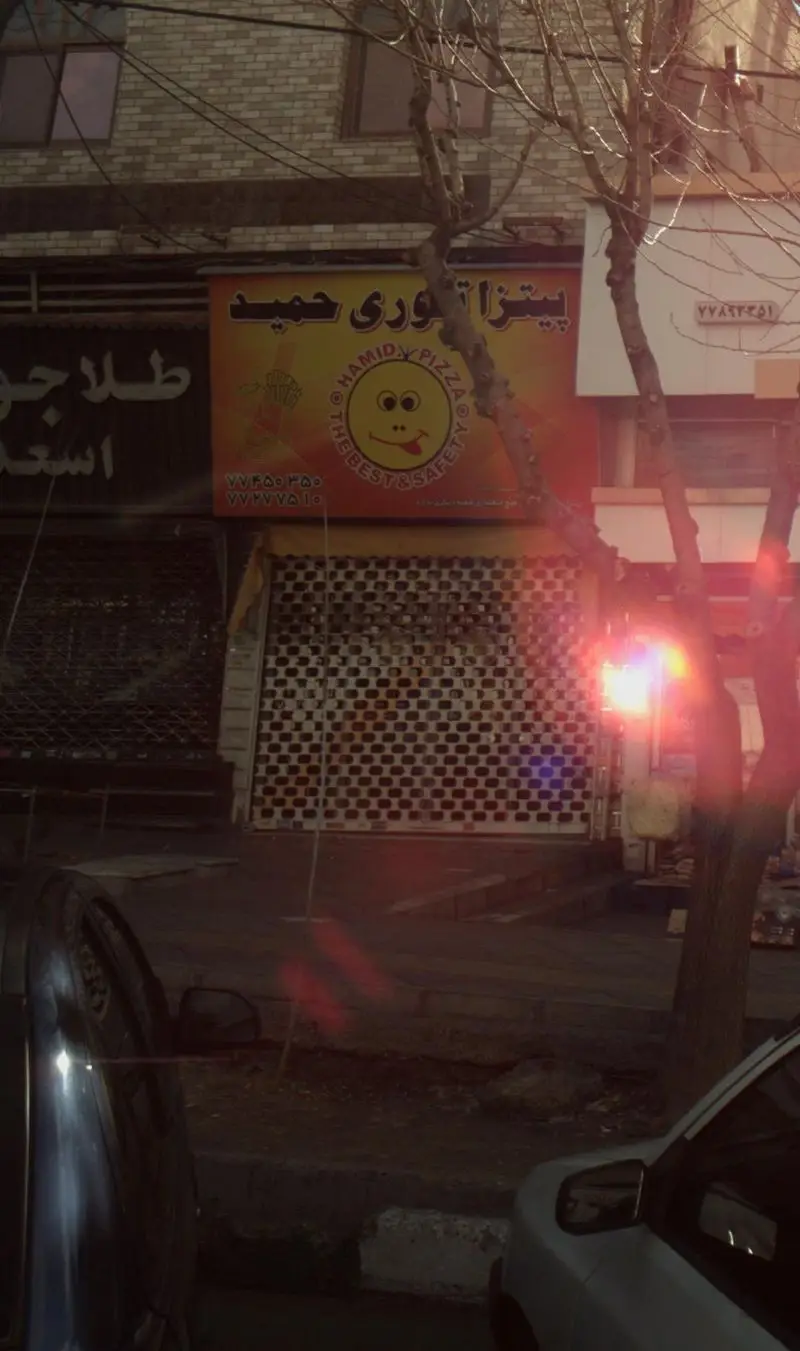 تابلوی زرد رنگ پیتزا تنوری حمید با لوگوی خندان و شماره تلفن در نمای جلوی مغازه بسته، همراه با درختان بی‌برگ و ماشین‌های پارک شده.