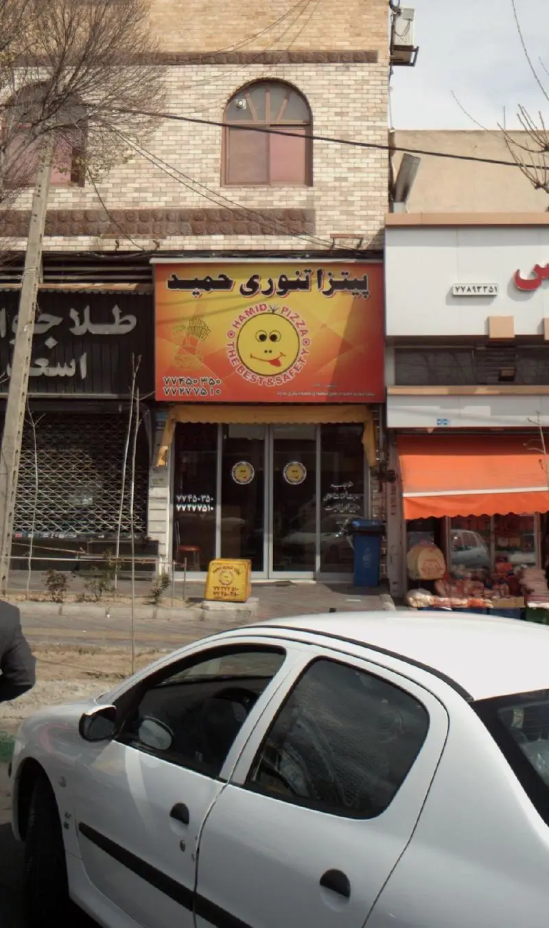 تابلوی بزرگ پیتزا تنوری حمید با لوگوی خندان و شماره تماس‌ها در نمای بیرونی رستوران، یک خودروی سفید در جلوی آن.
