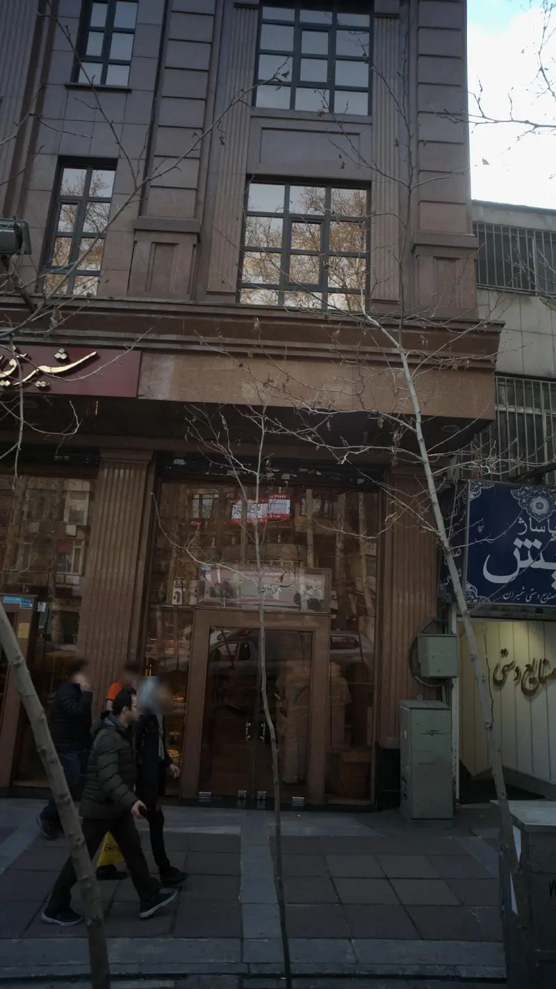 نمای بیرونی فروشگاه شیرینی لادن در یک روز آفتابی با پنجره های شیشه ای و بازتاب درختان خشکیده در خیابان شلوغ شهری تهران.