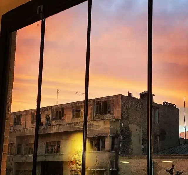 منظره دل‌انگیز غروب آسمان از پنجره کافه رستوران مستان؛ چشم‌اندازی زیبا با رنگ‌های گرم نارنجی و صورتی، رو به نمای شهری و یک ساختمان با دیوارنگاره جذاب.