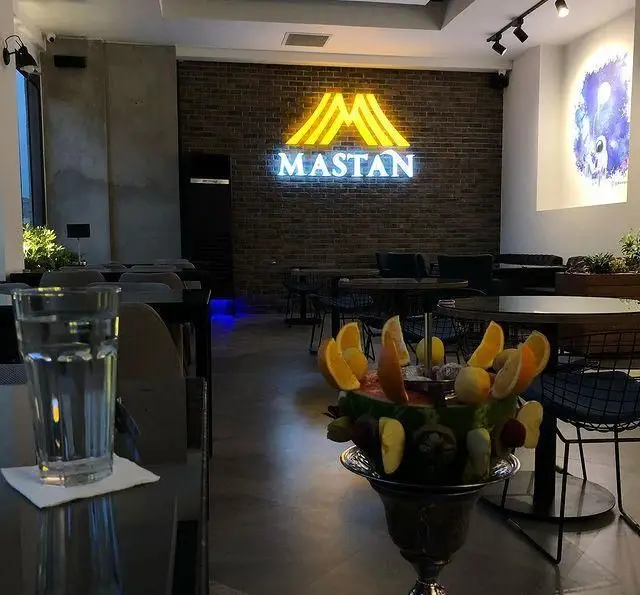 فضای داخلی کافه رستوران مستان با تابلوی نئونی بزرگ روی دیوار آجری، میزها و صندلی‌های مدرن، و قلیان میوه‌ای تزئین شده در پیش‌زمینه.