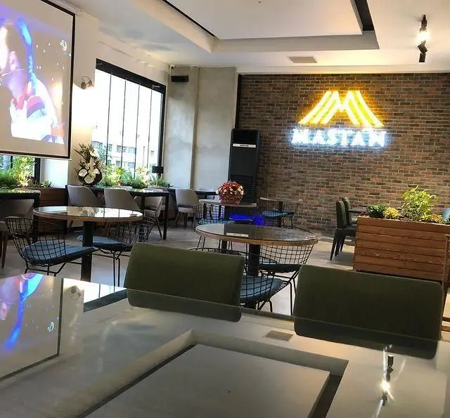 فضای داخلی دنج و مدرن کافه رستوران مستان با میز و صندلی‌های شیک، دیوار آجری، تابلوی نئون برند مستان و پروژکتور بزرگ.