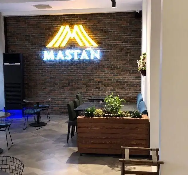 تصویری از فضای داخلی کافه رستوران مستان، با دیوار آجری، تابلو نئونی لوگوی M و نام "مستان" به رنگ آبی و زرد.