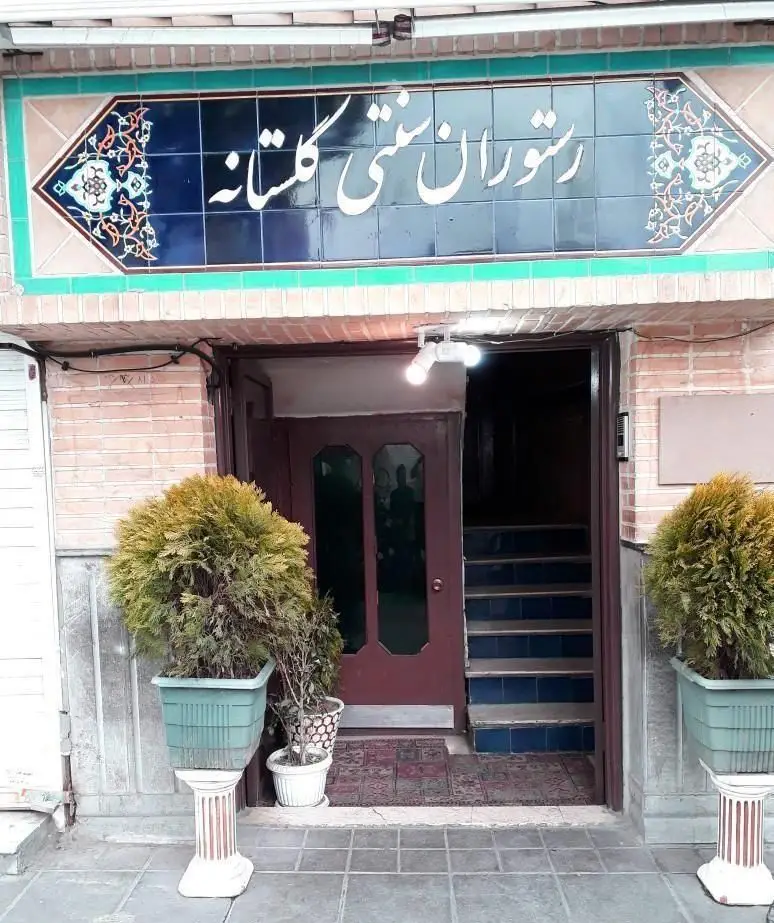 نمای ورودی رستوران سنتی گلستانه با تابلوی کاشی‌کاری شده آبی رنگ و خوشنویسی زیبا، گلدان‌های تزئینی و پله‌های ورودی.