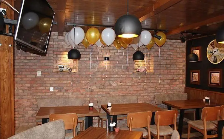فضای داخلی شیک و دلپذیر فست فود گاتا با دیوارهای آجری، میز و صندلی‌های چوبی، بالون‌های سفید و طلایی و نورپردازی گرم.