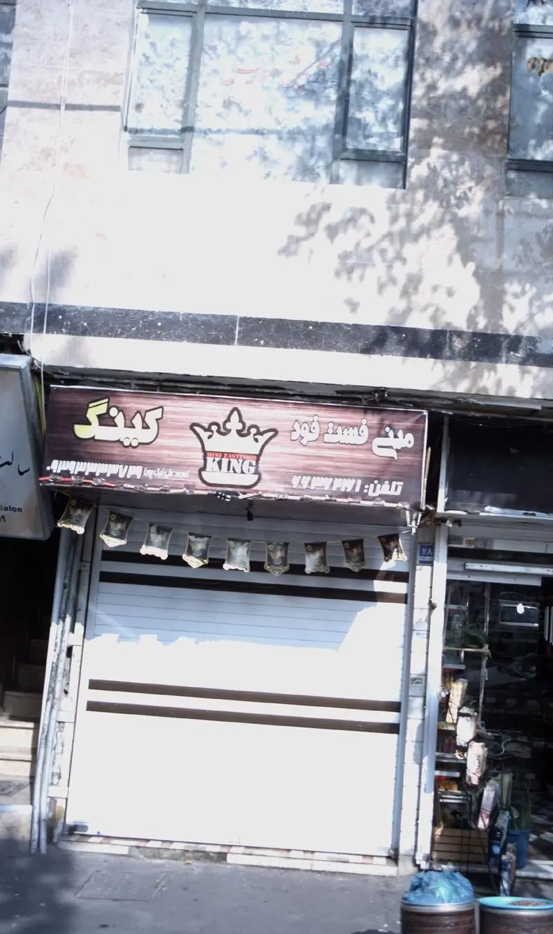 تصویری کامل از نمای بیرونی فست فود کینگ. تابلوی بزرگ قهوه‌ای رنگ با لوگوی تاج و نوشته "کینگ فست فود" بالای کرکره بسته مغازه به وضوح دیده می‌شود.