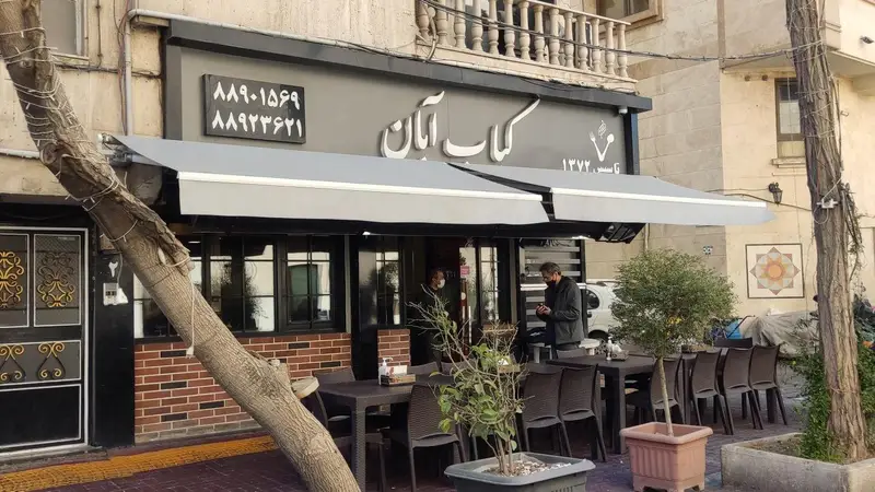 نمایی از رستوران کباب ابان با فضای نشیمن باز، آلاچیق‌های خاکستری و نمای آجری، همراه با شماره تلفن‌های ٨٨٩٠١٥٩٩ و ٨٨٩٢٣٦٢١.