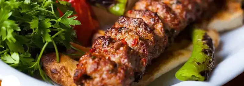 تصویری جذاب از کباب لذیذ و داغ آبان با گوشت چرخ‌کرده، به همراه فلفل کبابی و جعفری تازه، آماده سرو. بهترین کباب‌ها در کباب ابان.