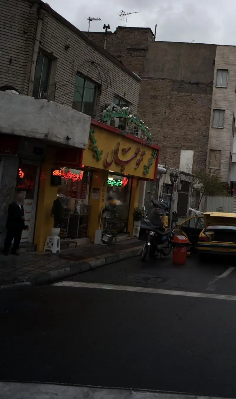 نمای پرجنب‌وجوش فروشگاه آبمیوه و فست فود عمو عباس در کنار خیابان، با تابلوی زرد رنگ و نورهای نئون در یک روز ابری شهر.