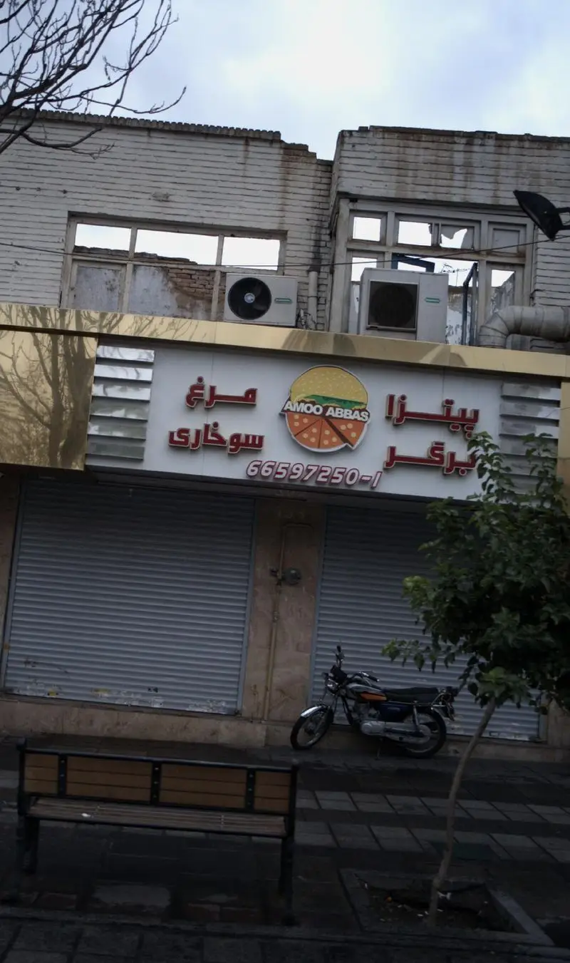 نمای بیرونی مغازه فست فود عمو عباس شامل تابلوی پیتزا، برگر و مرغ سوخاری در یک روز ابری، همراه با موتور و نیمکت.