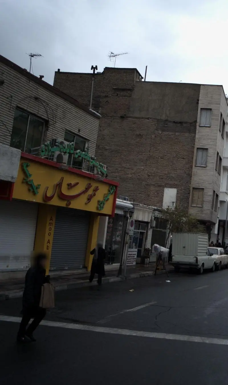 صحنه‌ای از یک خیابان شهری با تابلوی بزرگ و زرد رنگ مغازه عمو عباس، همراه با افراد در حال حرکت و ساختمان‌های آجری در پس‌زمینه.