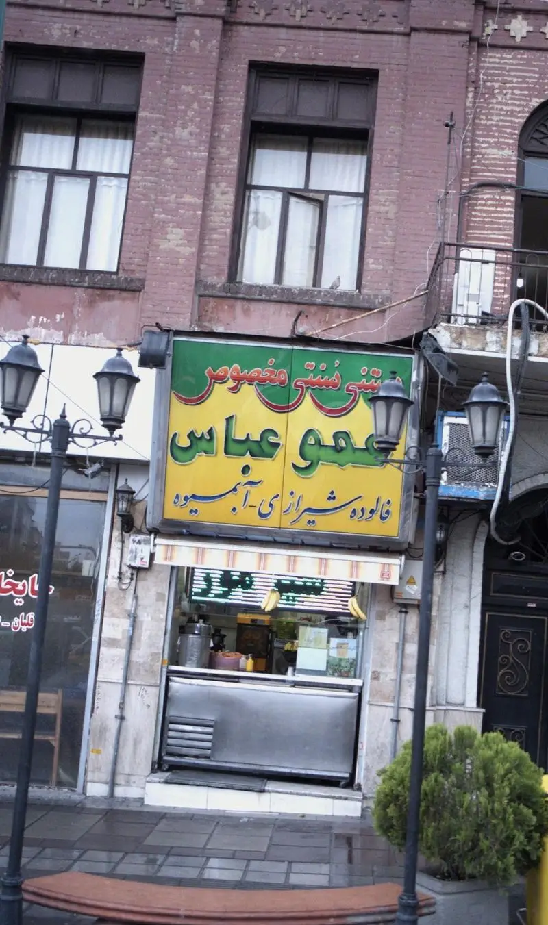 تصویری از فروشگاه عمو عباس، یک آبمیوه‌فروشی سنتی و ارائه‌دهنده فالوده شیرازی، با تابلو زردرنگ و ویترین استیل در خیابان.