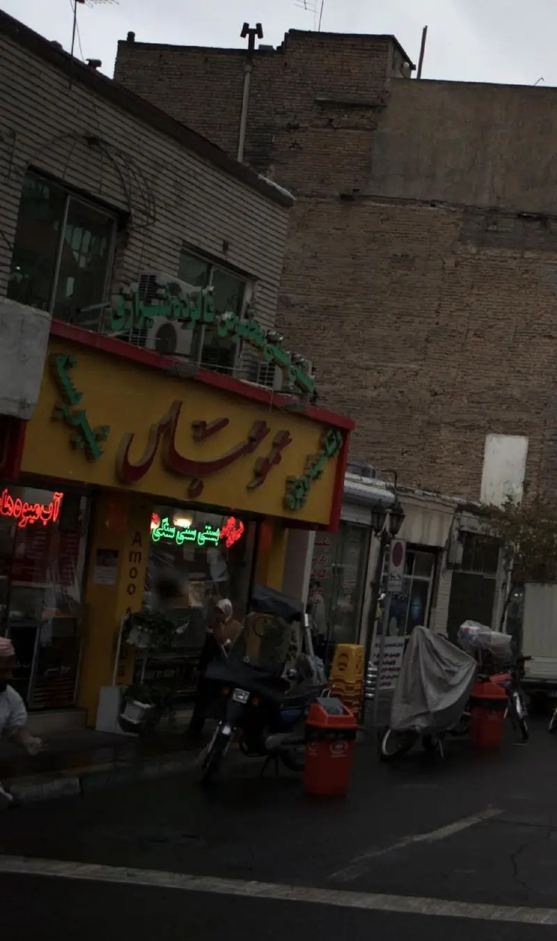 تصویر نمای خیابانی از کسب و کار بستنی و آبمیوه عمو عباس با تابلوی زرد رنگ و نوشته فارسی در یک روز ابری
