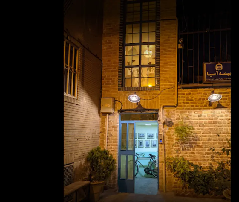 کافه کتاب نیکو: نمای شبانه فضایی دنج و دلنشین با معماری آجری، پنجره بلند نورانی، ورودی آبی و دوچرخه کلاسیک.