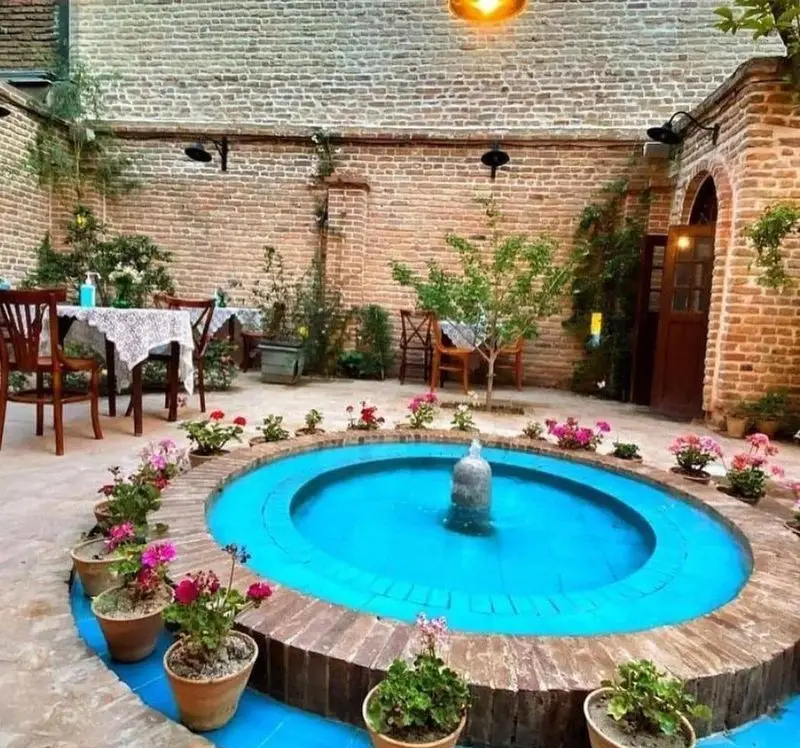 حیاط زیبای کافه کتاب نیکو با دیوارهای آجری، حوض آبی فواره‌دار، گلدان‌های گل و میز و صندلی‌های چوبی؛ محیطی آرام و دلنشین.