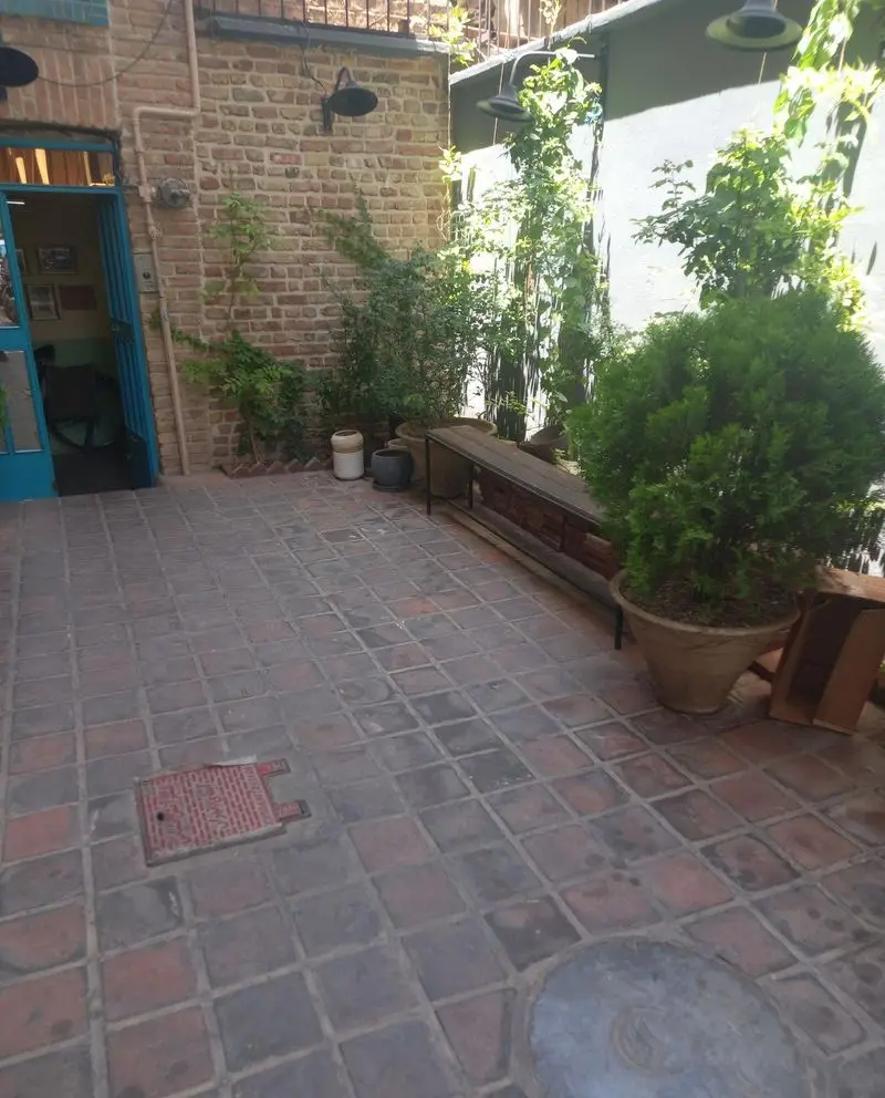 فضای باز کافه کتاب نیکو با دیوار آجری، درب آبی، گیاهان سبز و نیمکت چوبی، محیطی دنج و آرامش‌بخش برای مطالعه و دورهمی.