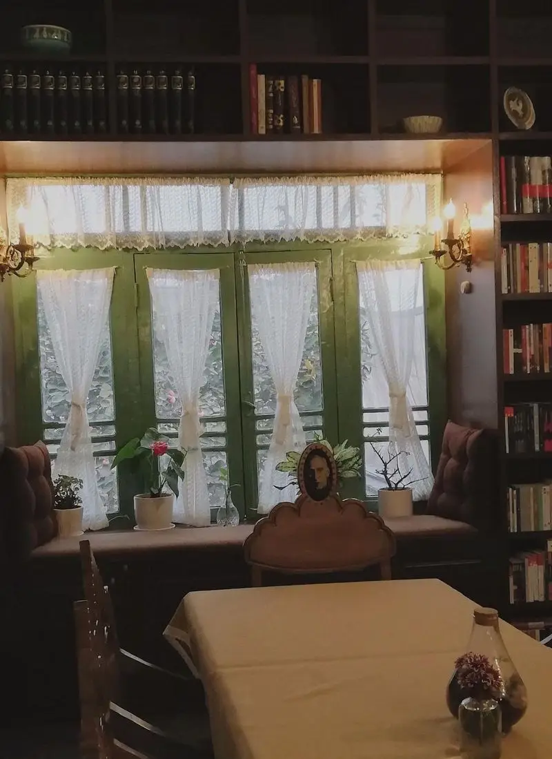 تصویری زیبا از کافه کتاب نیکو با فضای داخلی گرم، قفسه‌های کتاب، پنجره دلپذیر، پرده‌های توری و محیطی ایده‌آل برای مطالعه و آرامش.