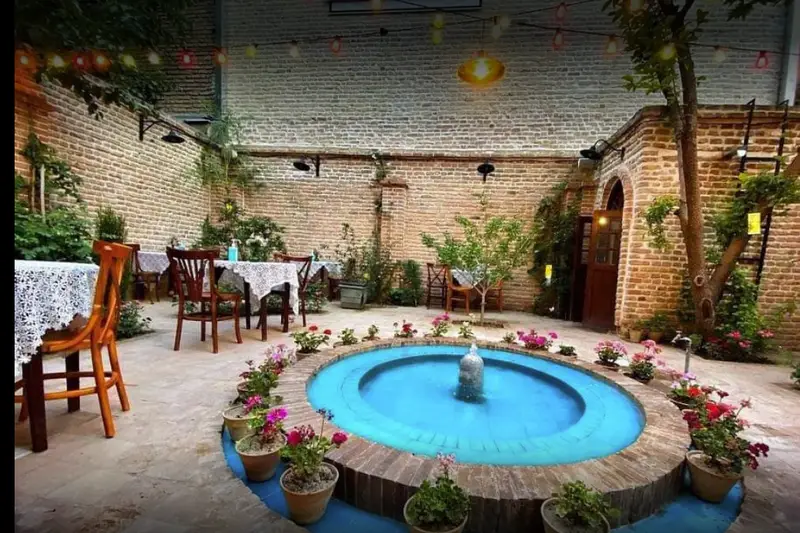 فضای دلنشین و سرسبز حیاط کافه کتاب نیکو با میز و صندلی‌های چوبی سفیدپوش، حوض آبنمای فیروزه‌ای، گلدان‌های گل رنگارنگ و دیوارهای آجری قدیمی و چراغ‌های تزئینی.