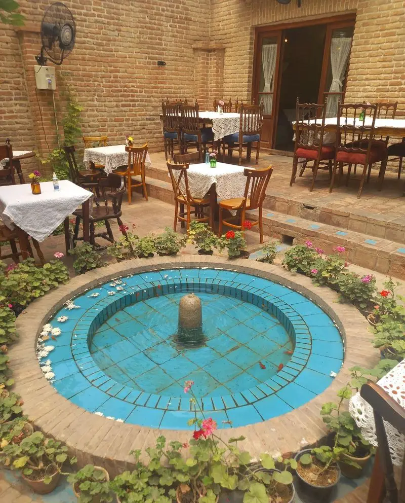 حیاط دلنشین کافه کتاب نیکو با فواره آبی رنگ مرکزی، میز و صندلی های چوبی و گلدان های گل زیبا در فضای آجری و سنتی.