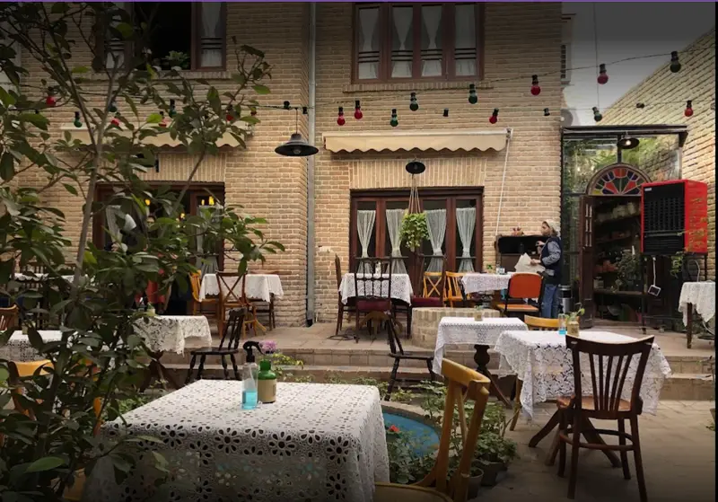 فضای دلنشین و دنج کافه کتاب نیکو در حیاطی سرسبز با میز و صندلی‌های چوبی و تزیینات نوری زیبا برای دورهمی و مطالعه.