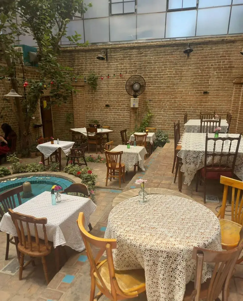 فضای باز دلنشین کافه کتاب نیکو: حیاطی زیبا با میز و صندلی‌های چوبی، آبنما، درخت و دیوار آجری، مکانی دنج برای مطالعه و گفتگو.