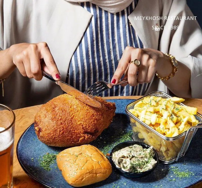 تصویری از مشتری در رستوران میخوش در حال برش شنیسل مرغ سوخاری یا چیکن کوردن بلو با سیب زمینی، نان و سس قارچ روی میز.