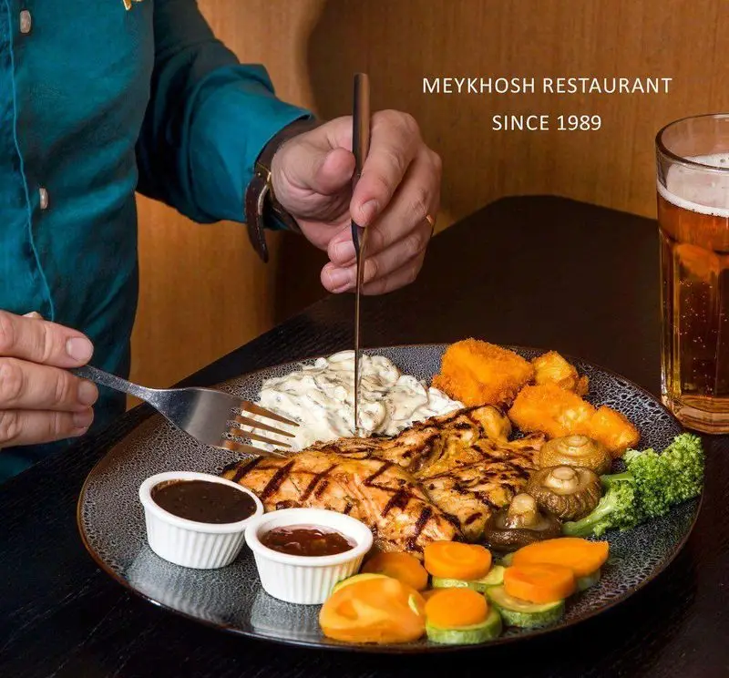 شخصی در حال برش استیک مرغ گریل شده با سس قارچ و سبزیجات متنوع در رستوران میخوش. تجربه‌ای لذیذ از غذای اصلی در محیطی دلنشین.
